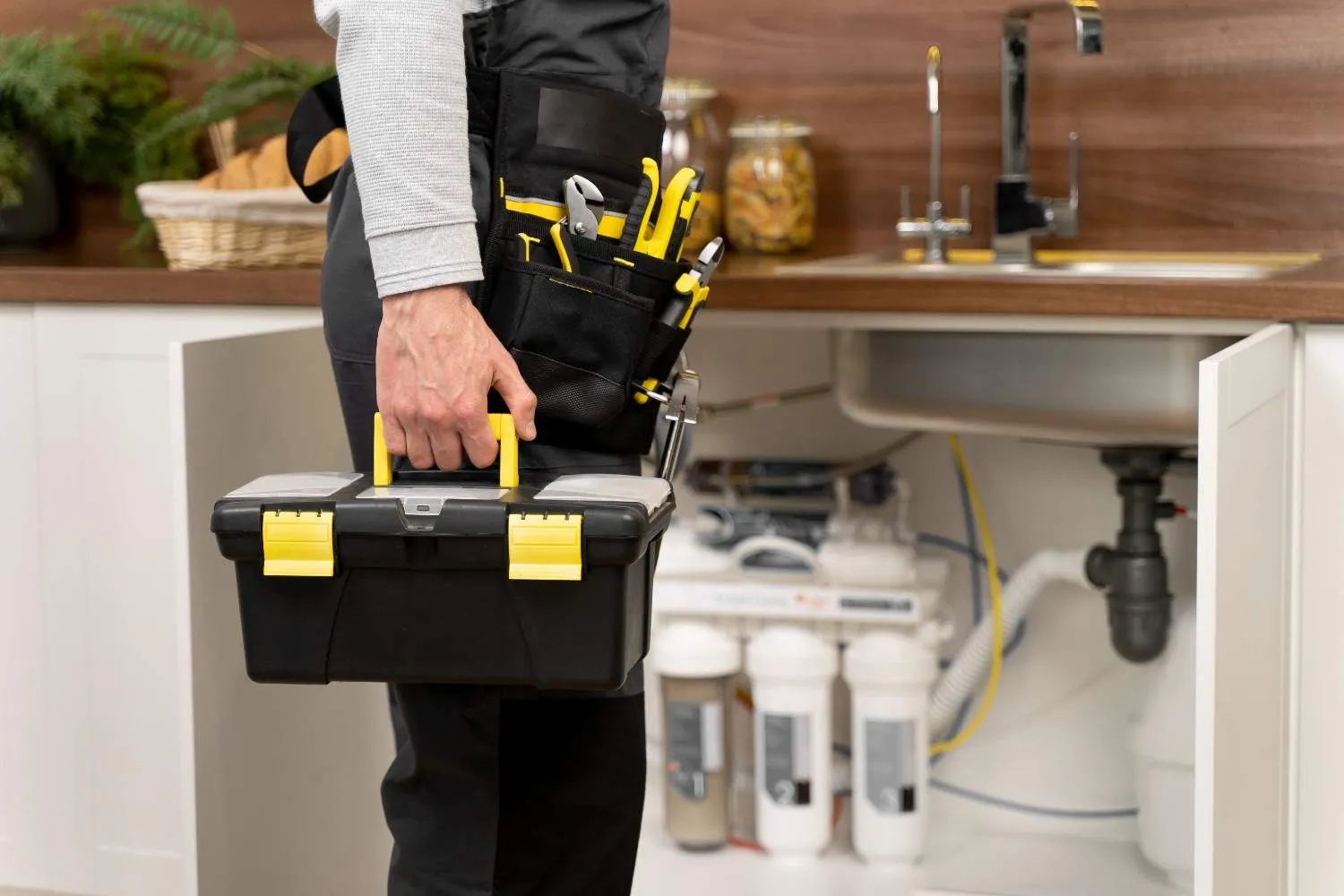 Plumber with a toolbox and tool belt standing by an open kitchen sink cabinet