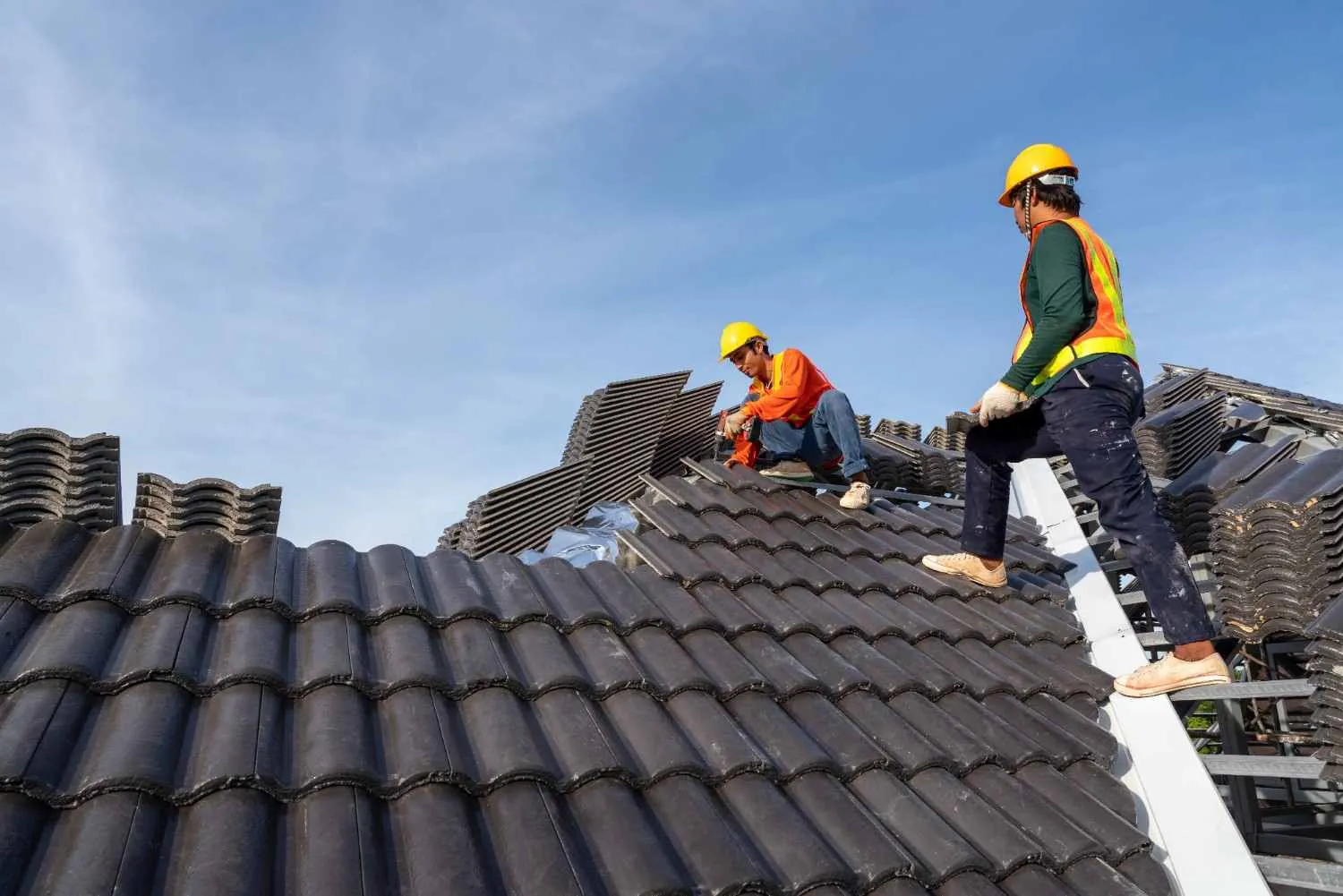 Two roofers in hard hats laying dark tiles on a house roof